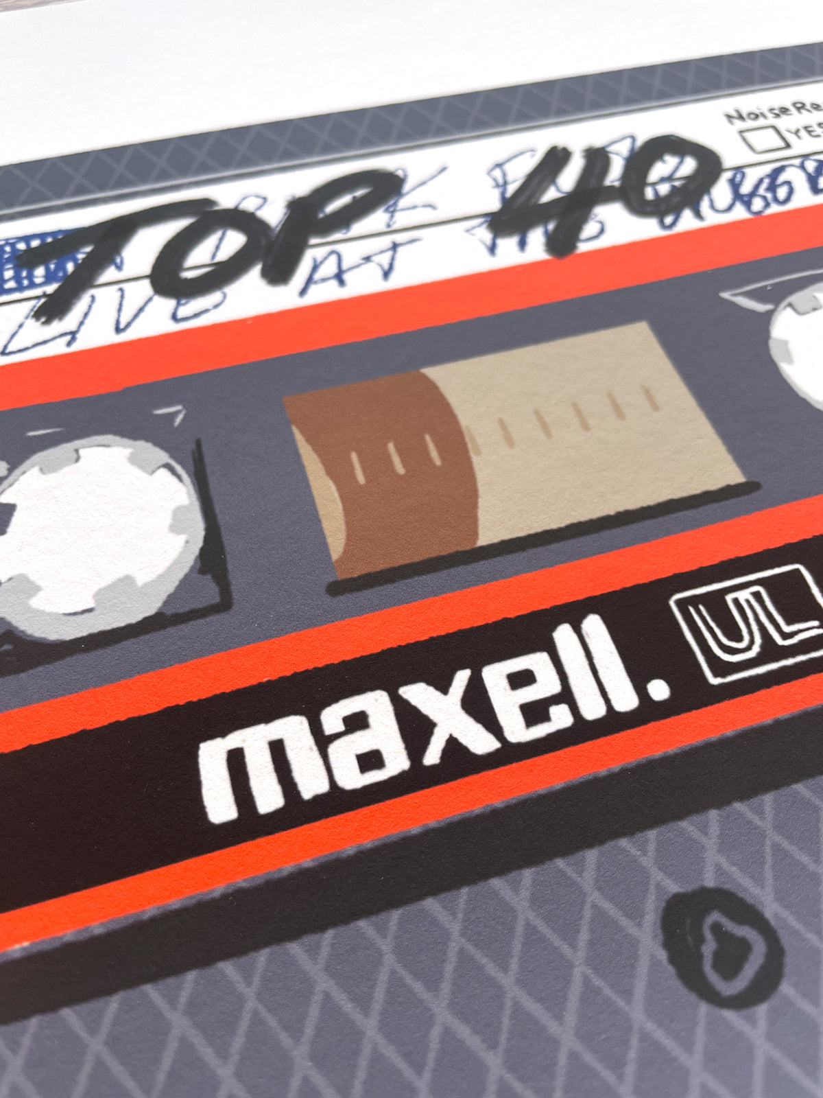 A close-up of a cassette tape graphic print featuring the brand name 'maxell.' in white on a black band with a bright orange stripe, alongside a beige window area and gray outer casing.