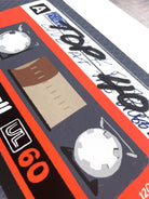 A close-up of a retro cassette tape illustration or print, featuring orange-red borders, a dark gray body, beige inner tape window, and stylized text including 'Top 40' and '60'.