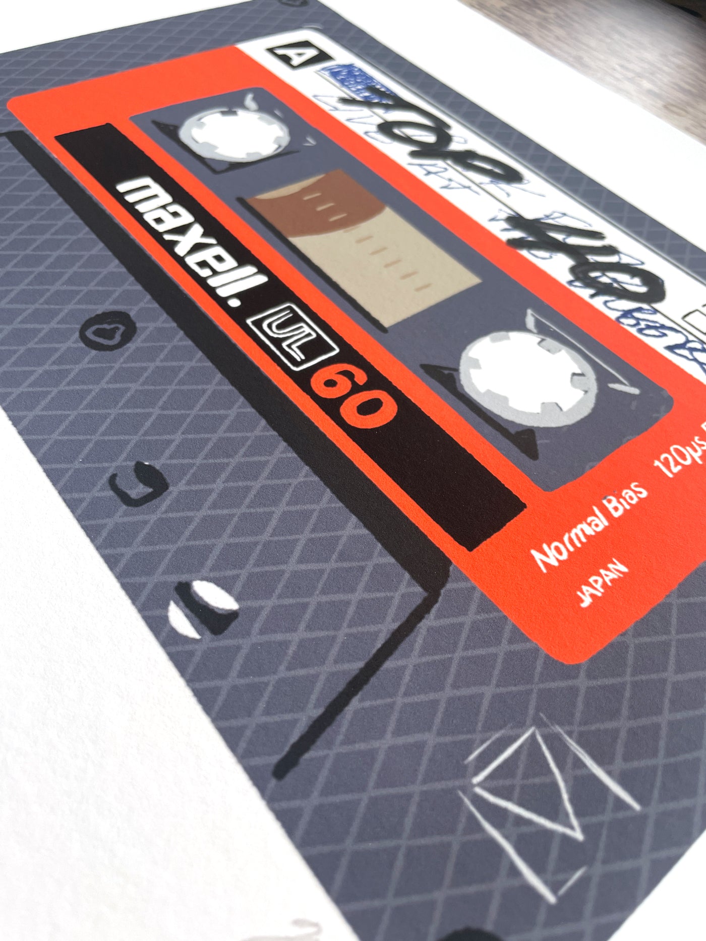 A close-up of a printed cassette tape illustration or poster featuring a gray patterned background with a large orange and black Maxell UC-60 label.