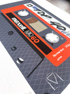 A close-up of a printed cassette tape illustration or poster featuring a gray patterned background with a large orange and black Maxell UC-60 label.