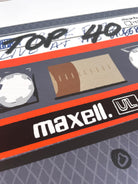 A close-up of a cassette tape graphic print featuring the brand name 'maxell.' in white on a black band with a bright orange stripe, alongside a beige window area and gray outer casing.